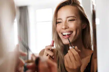 Lächelnde Frau trägt Lipgloss vor dem Spiegel auf – Beauty-Routine mit guter Laune