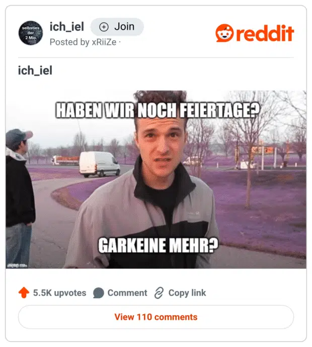 Ein junger Mann steht fragend auf einem Parkplatz mit lila eingefärbtem Boden. Im Bild steht in Großbuchstaben: „HABEN WIR NOCH FEIERTAGE?“ und darunter: „GARKEINE MEHR?“ Im Hintergrund sind weitere Personen, Autos und kahle Bäume zu sehen.
