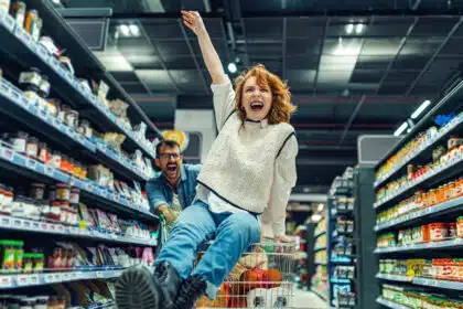 Junge Frau sitzt lachend in einem Einkaufswagen, während ein Mann sie durch einen Supermarkt schiebt – lebhafte Szene mit Spaß beim Einkaufen, ideal als Vorschau-Bild für humorvolle Social-Media-Posts oder Beiträge über Paar-Alltag und Lebensfreude auf Twitterperlen.de.