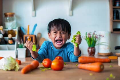 Wütender kleiner Junge sitzt am Küchentisch und schreit, während er mit beiden Händen Salat zerreißt – frustriertes Kind umgeben von frischem Gemüse wie Tomaten und Karotten, Symbolbild für Essensverweigerung oder Familienalltag mit Kindern auf Twitterperlen.de.
