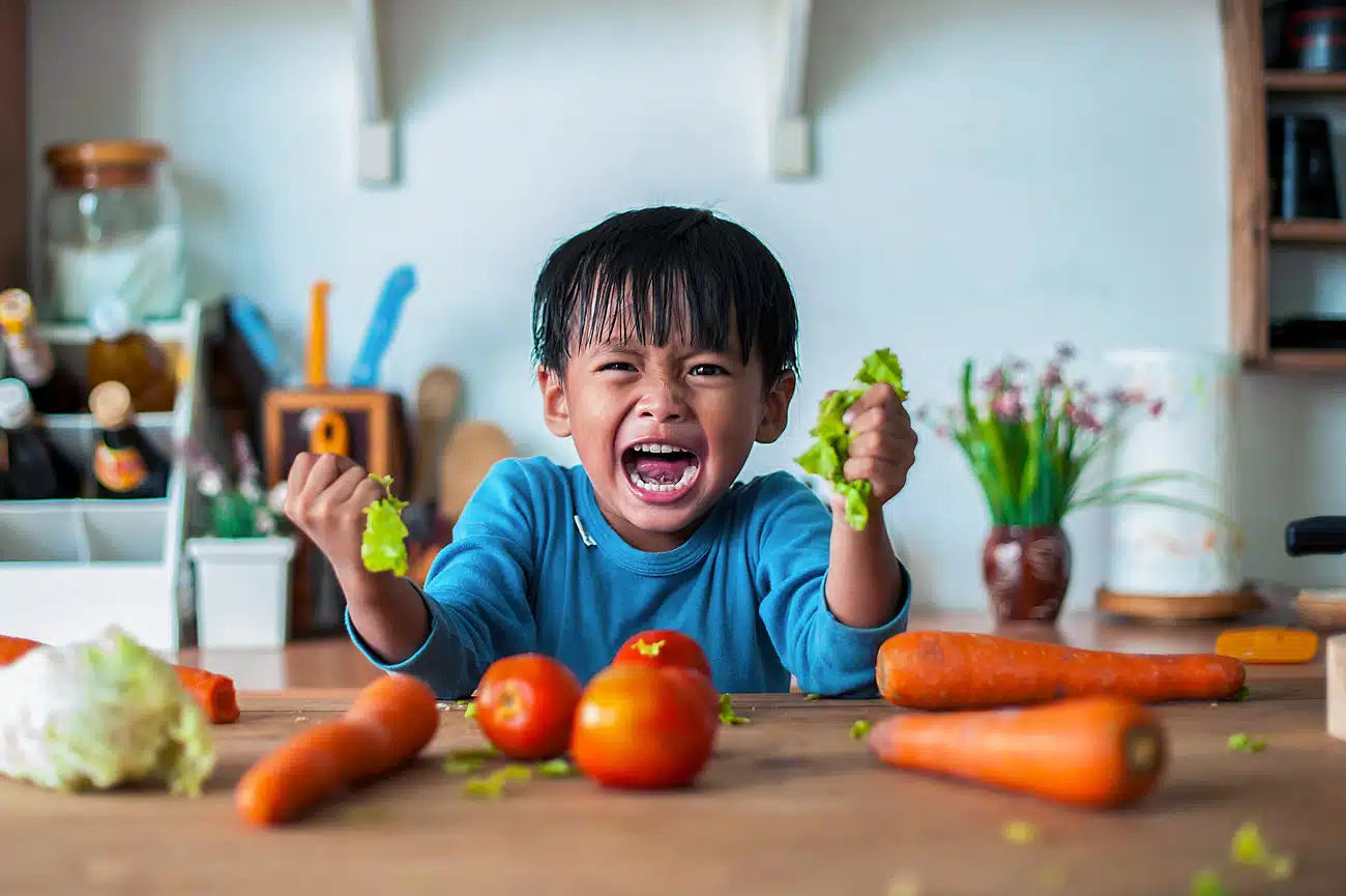 Wütender kleiner Junge sitzt am Küchentisch und schreit, während er mit beiden Händen Salat zerreißt – frustriertes Kind umgeben von frischem Gemüse wie Tomaten und Karotten, Symbolbild für Essensverweigerung oder Familienalltag mit Kindern auf Twitterperlen.de.
