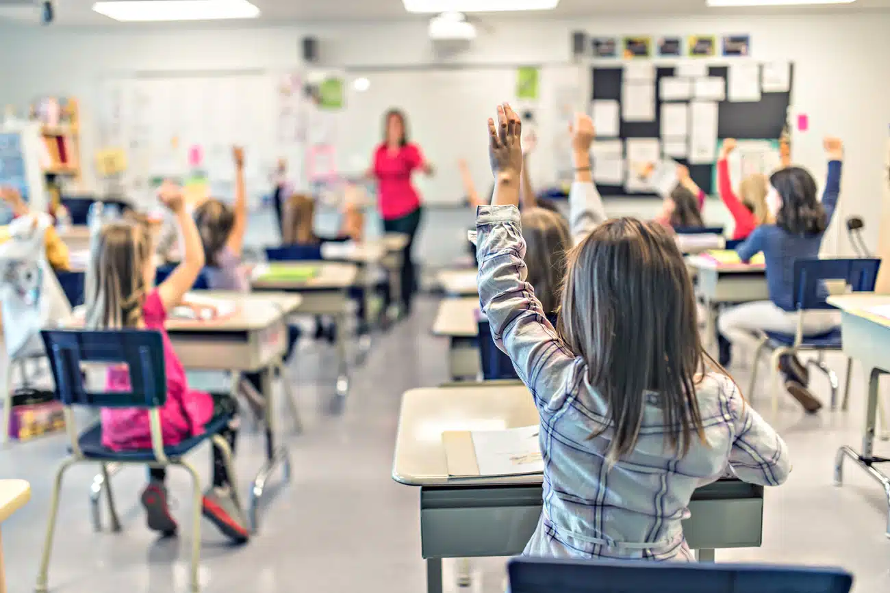 Grundschulkinder im Klassenzimmer heben motiviert die Hand, um sich am Unterricht zu beteiligen – lebendiger Schulalltag mit engagierter Lehrerin im Hintergrund, Fokus auf Bildung, Beteiligung und moderne Lernumgebung