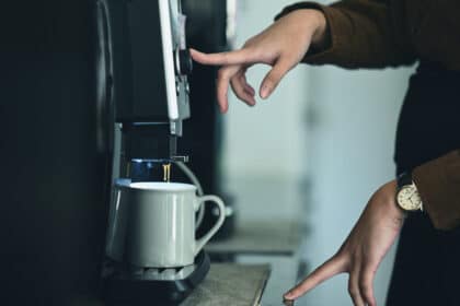 Person mit Armbanduhr bedient Kaffeemaschine im Büro – frischer Kaffee tropft in eine weiße Tasse