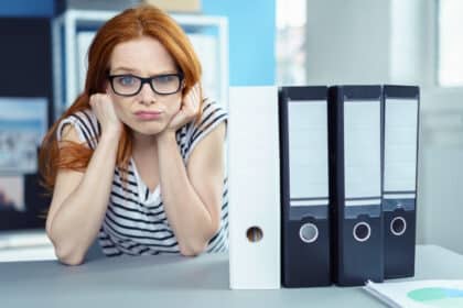 Unmotivierte junge Frau mit Brille schaut genervt auf Aktenordner im Büro – Symbolbild für Motivationslosigkeit, Frust, Demotivation oder Bürokratie.