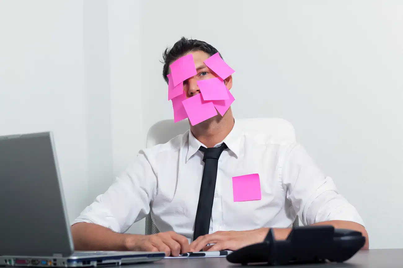 Ein Büroangestellter sitzt erschöpft an seinem Schreibtisch, bekleidet mit weißem Hemd und schwarzer Krawatte. Sein Gesicht ist fast vollständig mit pinken Haftnotizzetteln bedeckt, einer klebt zusätzlich auf seiner Brust. Vor ihm stehen ein aufgeklappter Laptop und ein Tischtelefon. Die Szene wirkt humorvoll überzeichnet und symbolisiert Überforderung oder Bürofrust.