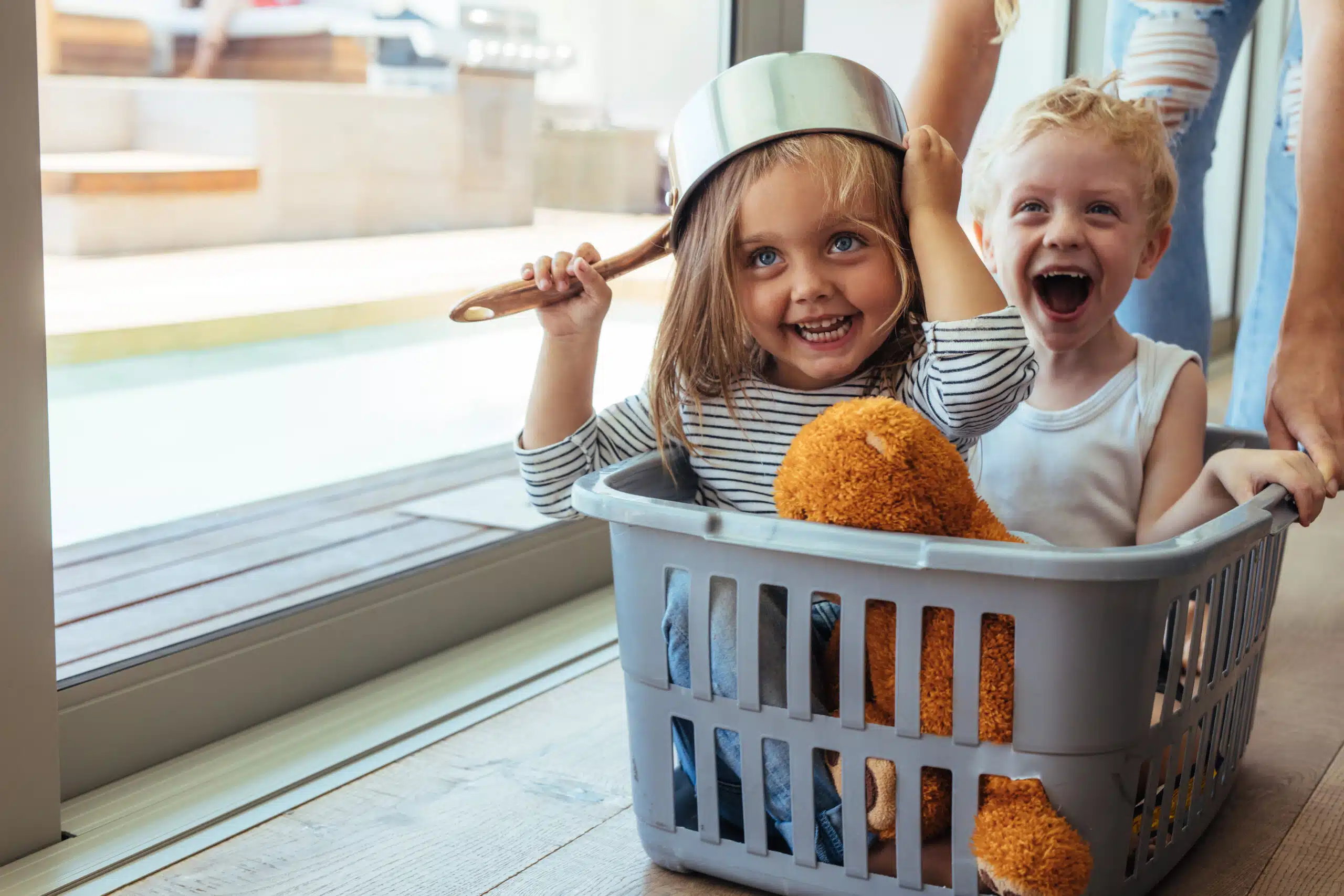 Kinder spielen fantasievoll mit den Eltern und Alltagsgegenständen – Kochtopf als Helm und Wäschekorb als Spielgefährt