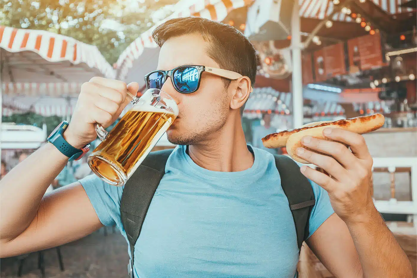 Deutscher Mann mit Sonnenbrille genießt draußen ein großes Glas Bier und eine deutsche Bratwurst in einem Biergarten.