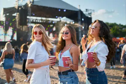 Drei junge Frauen stehen vor einer Bühne auf einem Festival und tanzen lachend zusammen.