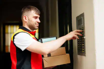 Postbote von DHL klingelt an einer Haustür und hält mehrere Pakete in der Hand.