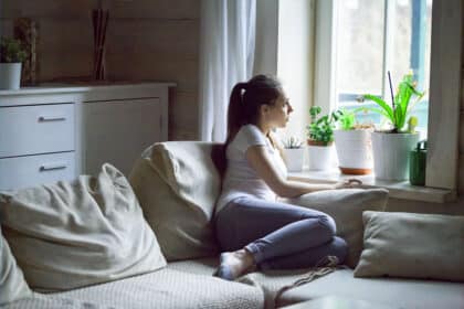 Junge Frau sitzt nachdenklich auf dem Sofa und blickt aus dem Fenster, sie scheint recht introvertiert zu leben.