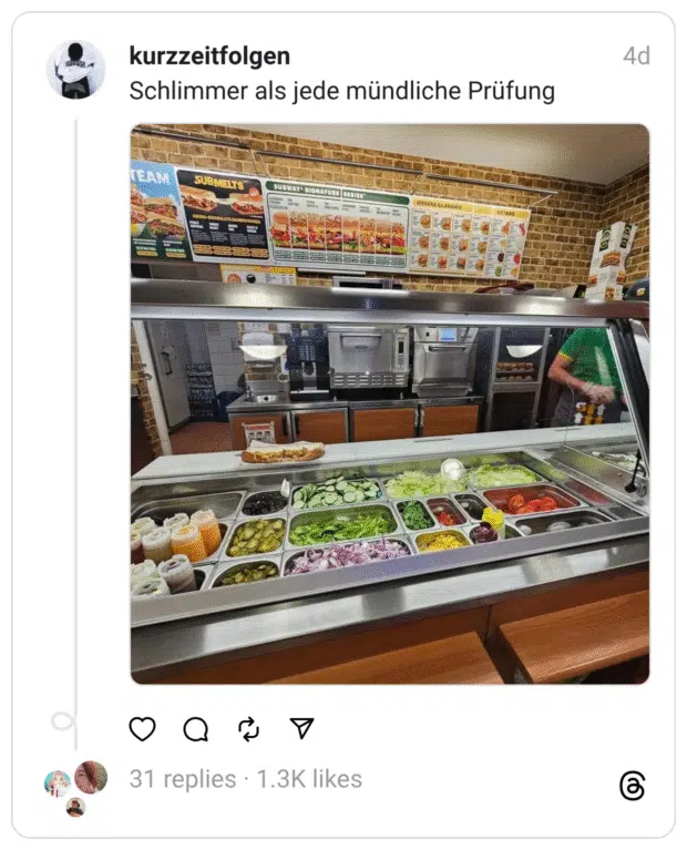 Innenaufnahme eines Subway-Restaurants, aufgenommen aus Kundensicht an der Theke. Hinter der Glasvitrine sind zahlreiche Zutaten wie Salat, Tomaten, Gurken, Zwiebeln, Mais, Soßenflaschen und weitere Toppings sauber aufgereiht. Ein Mitarbeiter in grünem Shirt steht bereit. Die Bildunterschrift lautet: „Schlimmer als jede mündliche Prüfung“ – eine humorvolle Anspielung auf die Überforderung, sich spontan für Zutaten beim Subway-Bestellen entscheiden zu müssen.