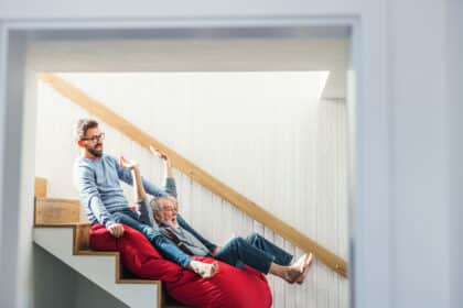 Junger Mann und älterer Mann rutschen lachend gemeinsam auf einem Sitzsack im Treppenhaus die Treppe hinunter.