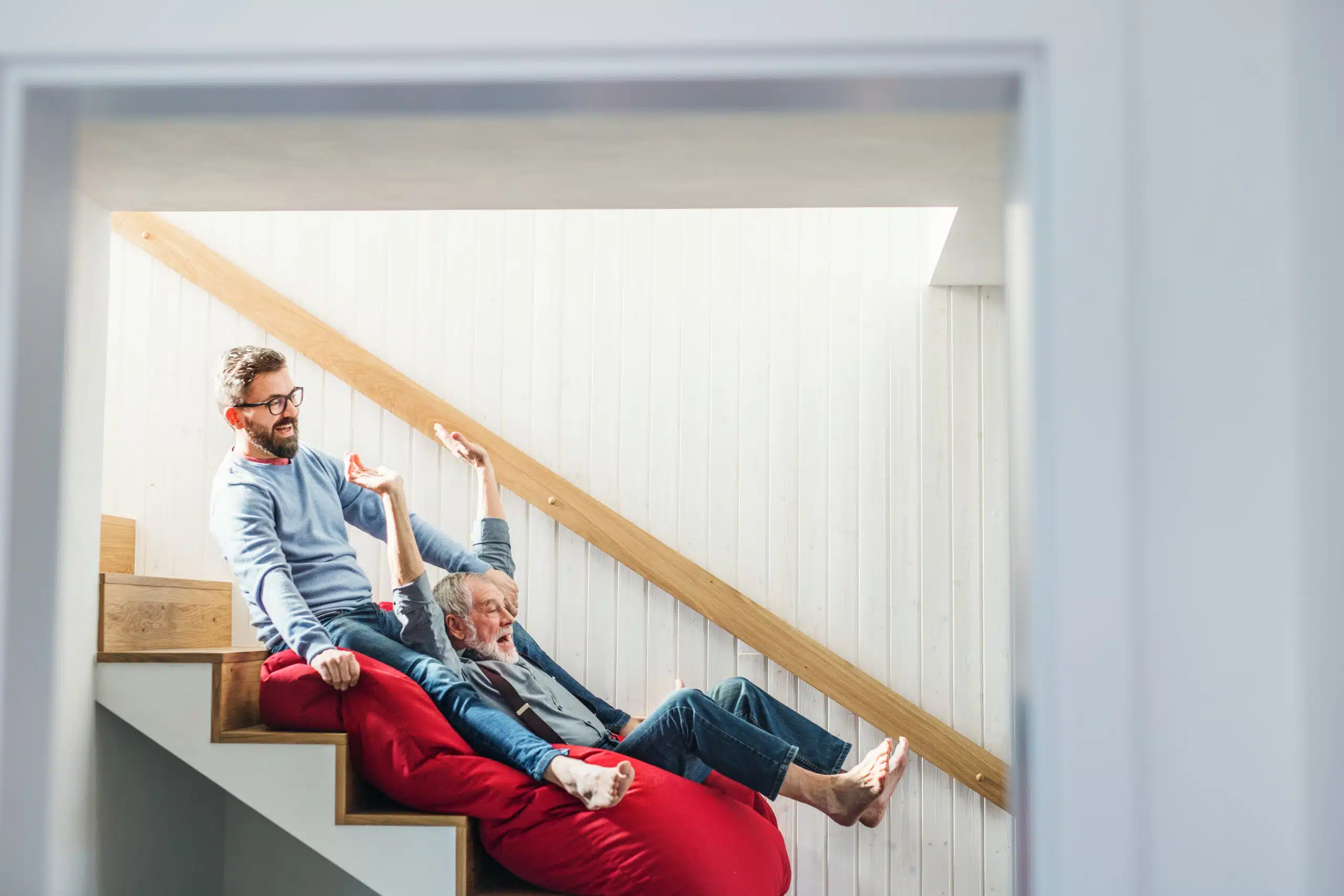 Junger Mann und älterer Mann rutschen lachend gemeinsam auf einem Sitzsack im Treppenhaus die Treppe hinunter.