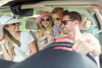 Glückliche Familie mit zwei Kindern trägt Sonnenbrillen und lacht gemeinsam auf einer Autofahrt in den Urlaub.
