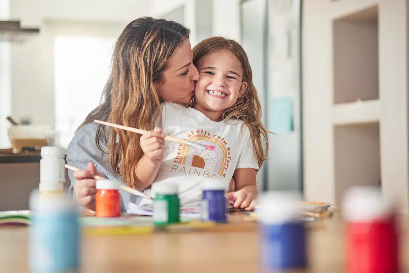 Mutter küsst lachende Tochter beim Malen mit bunten Farben.