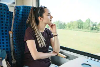 Junge Frau sitzt am Fenster eines Zuges und blickt nachdenklich in die vorbeiziehende Landschaft