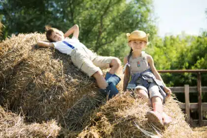 Zwei Kinder in Latzhosen und Gummistiefeln spielen fröhlich auf einem Heuhaufen im Dorf an einem sonnigen Sommertag – idyllische Dorfkindheit mit Naturerlebnis.