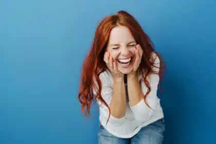 Junge Frau mit roten Haaren lacht herzlich mit geschlossenen Augen und Händen im Gesicht vor blauer Wand – Ausdruck von Freude, Überraschung oder Euphorie.