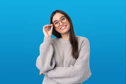 Junge Frau mit Brille und grauem Pullover lächelt vor blauem Hintergrund.