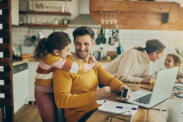 Glückliche Eltern mit Kindern: Vater arbeitet im Homeoffice mit Laptop, während Tochter ihn umarmt und Mutter mit zweitem Kind am Küchentisch lacht.
