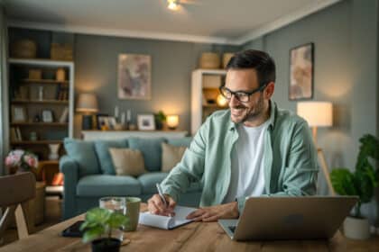 Erwachsener Mann arbeitet glücklich zu Hause am Schreibtisch mit Laptop und Notizbuch.