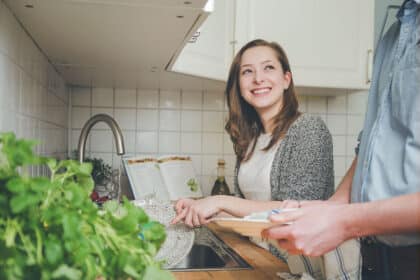 Junge Frau spült Geschirr in der Küche und lächelt eine zweite Person an, die beim Abtrocknen hilft.