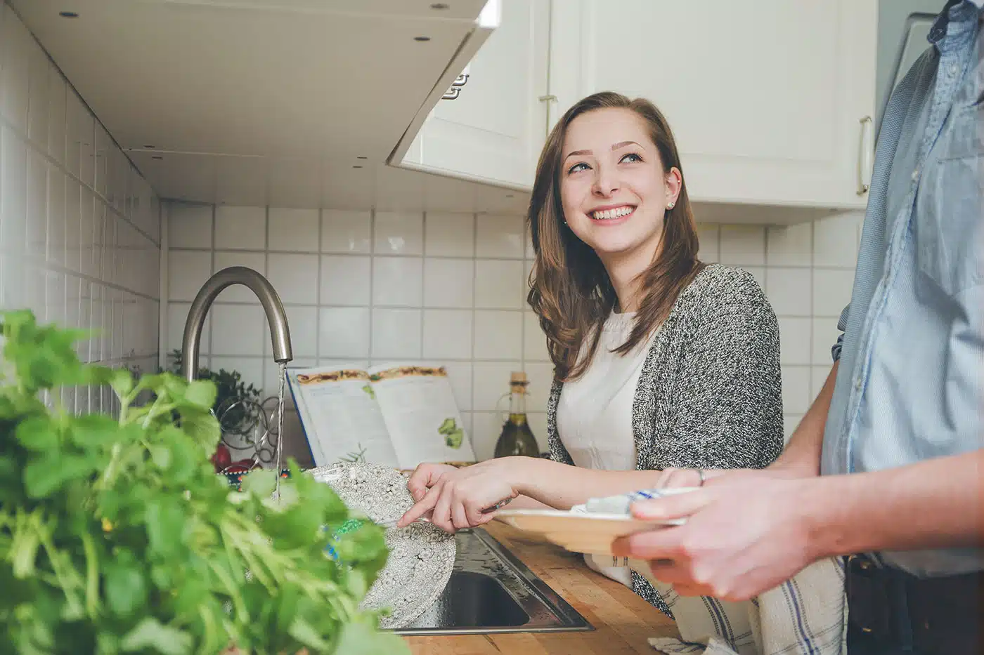 Junge Frau spült Geschirr in der Küche und lächelt eine zweite Person an, die beim Abtrocknen hilft.