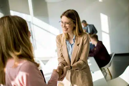 Junge Frau mit Brille gibt einer anderen Person im Büro die Hand und lächelt freundlich, um ja zu sagen.