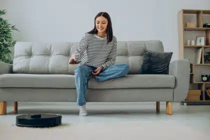 Junge Frau sitzt lächelnd auf einem Sofa und steuert mit einer Fernbedienung einen Saugroboter im Wohnzimmer.