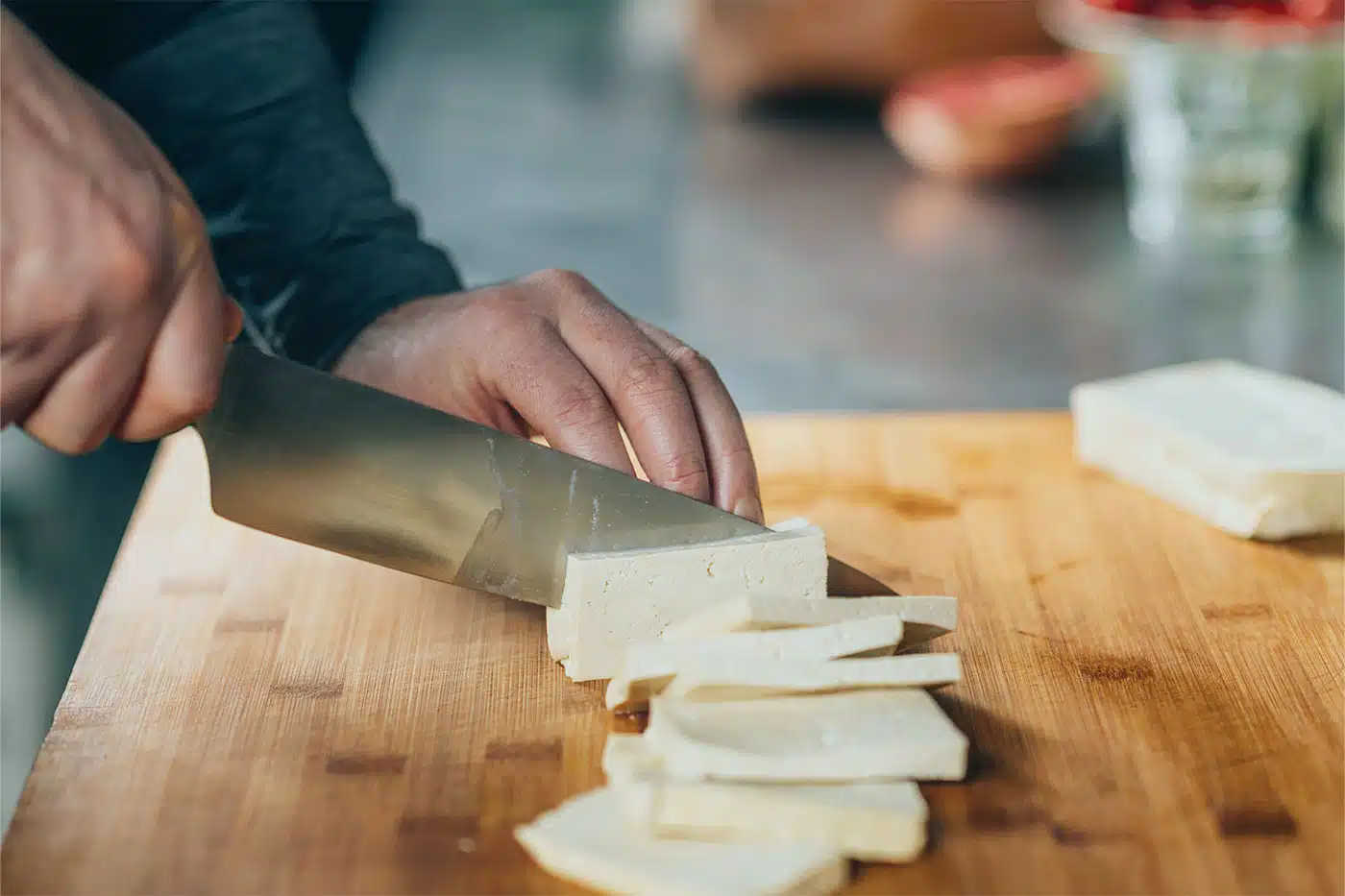Nahaufnahme einer Hand, die frischen Tofu auf einem Holzbrett in Scheiben schneidet.
