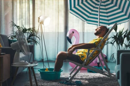 Mann entspannt im Wohnzimmer auf einem Liegestuhl unter einem Sonnenschirm, mit Füßen in einer Wasserwanne, umgeben von Sommerdeko wie einem aufblasbaren Flamingo und einer Gummiente – kreative Abkühlung bei Sommerhitze zu Hause.