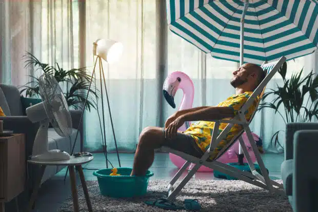 Mann entspannt im Wohnzimmer auf einem Liegestuhl unter einem Sonnenschirm, mit Füßen in einer Wasserwanne, umgeben von Sommerdeko wie einem aufblasbaren Flamingo und einer Gummiente – kreative Abkühlung bei Sommerhitze zu Hause.