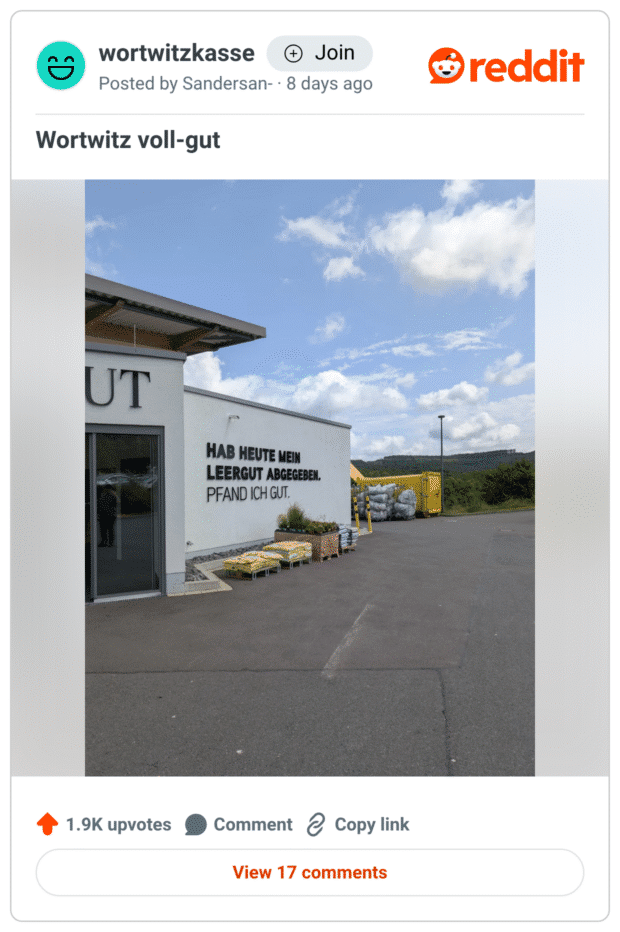 Foto einer Gebäudewand auf einem Hof. Auf der weißen Fassade steht in großen schwarzen Lettern: „HAB HEUTE MEIN LEERGUT ABGEGEBEN. PFAND ICH GUT.“ Davor Paletten mit Säcken und Pflanzen, rechts im Hintergrund ein gelber Container. Wortspiel mit „Pfand“ statt „fand“.