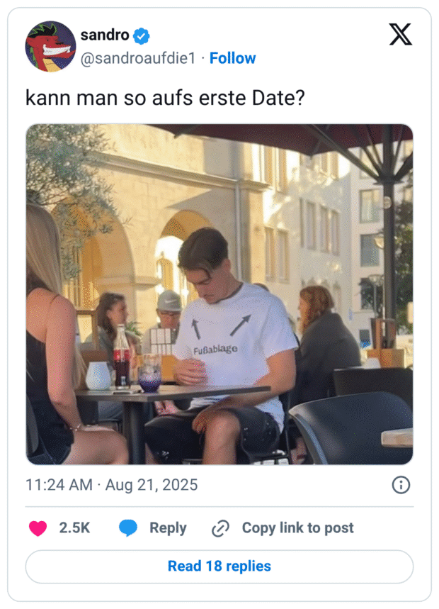 Tweet „kann man so aufs erste Date?“. Foto aus einem Straßencafé: Ein junger Mann sitzt einer Frau gegenüber. Er trägt ein weißes T-Shirt mit dem Aufdruck „Fußablage“ und zwei Pfeilen darüber.