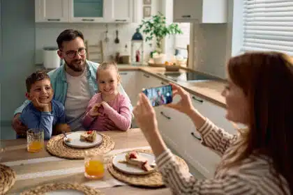 Eine Mutter macht mit ihrem Smartphone ein Bild von ihrem lachenden Mann und ihren zwei Kindern, die glücklich an einem Küchentisch sitzen.