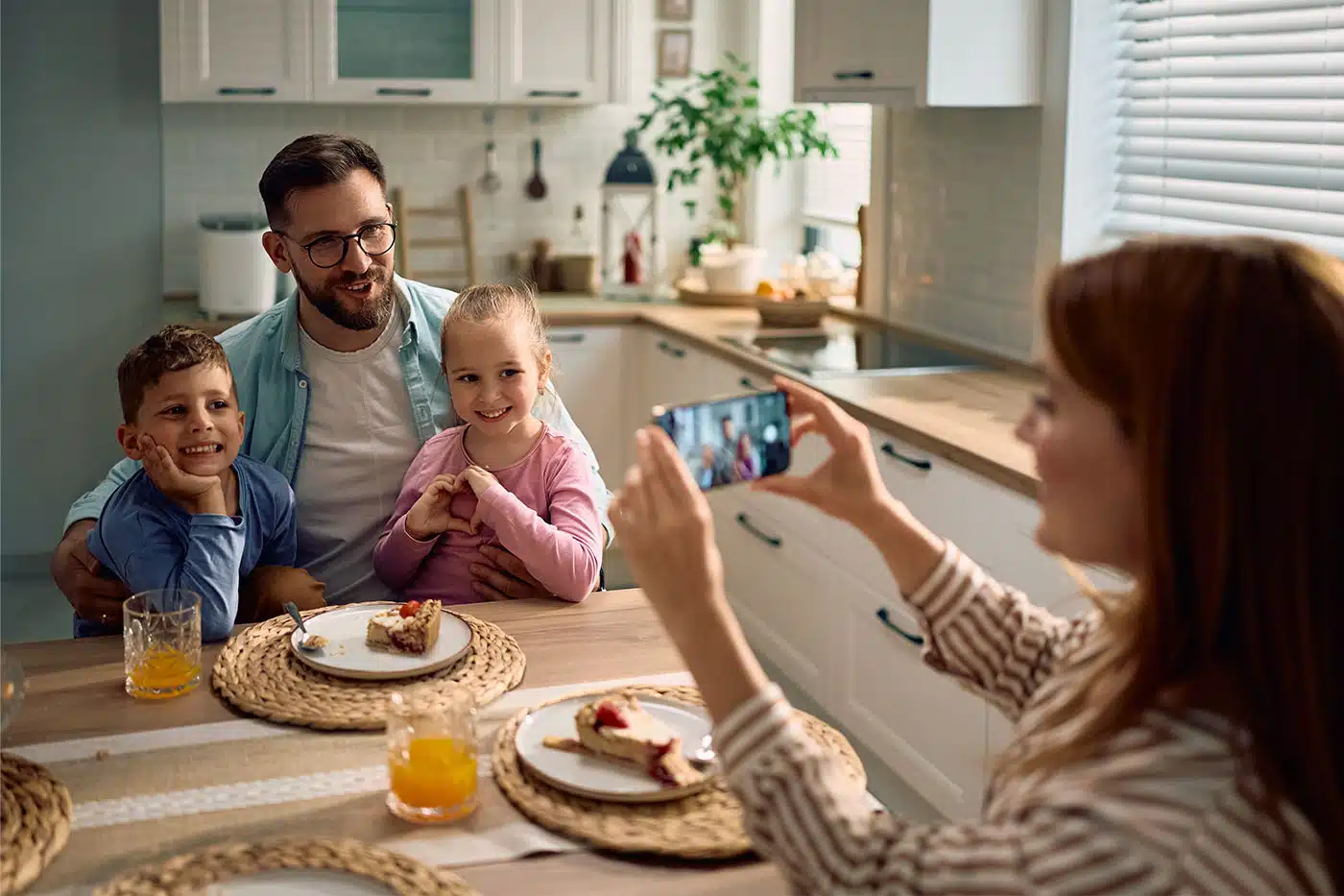 Eine Mutter macht mit ihrem Smartphone ein Bild von ihrem lachenden Mann und ihren zwei Kindern, die glücklich an einem Küchentisch sitzen.