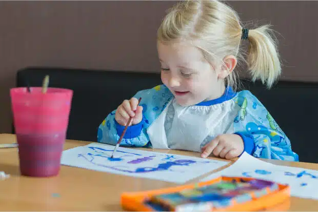 Ein kleines blondes Mädchen mit einer Malschürze sitzt glücklich und konzentriert an einem Tisch und malt mit blauer Farbe ein Bild für ihre Eltern.