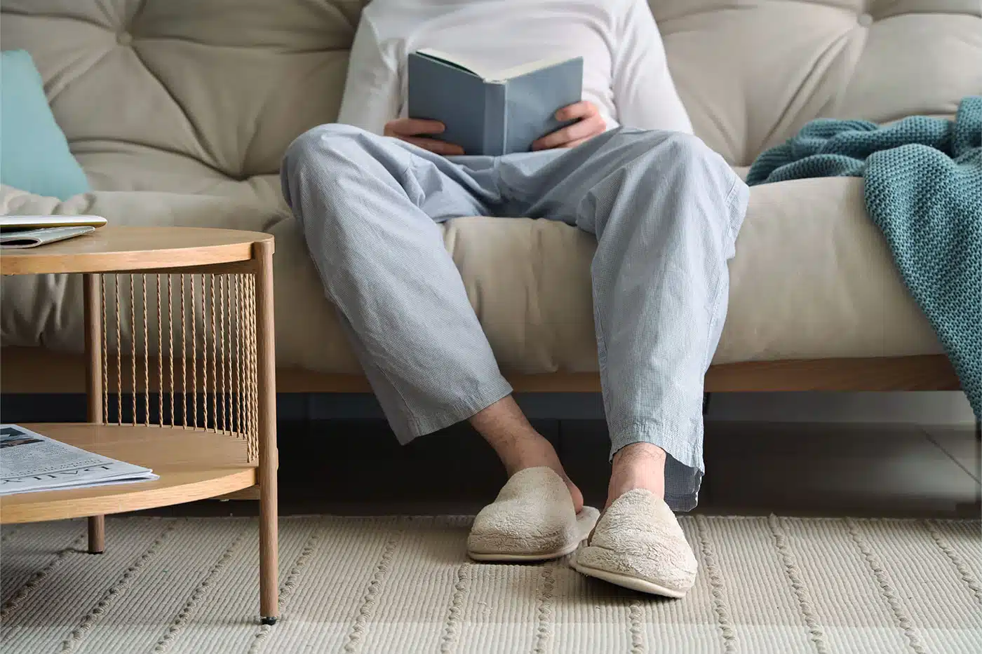 Eine Person in bequemer Kleidung und Hausschuhen sitzt gemütlich auf einem Sofa und liest ein Buch.