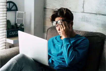 Ein junger Mann mit Brille sitzt erschöpft auf einem Sofa und hält sich die Hand an die Stirn, während er auf Jobsuche ist.