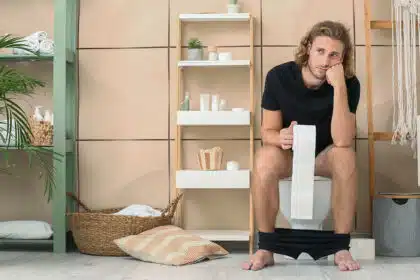Ein junger Mann mit sitzt auf dem Klo und hält eine Rolle Toilettenpapier in der Hand.