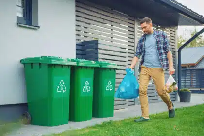Ein Mann im Freien bringt Müll und Wertstoffe in Beuteln zu den drei grünen Mülltonnen vor seinem Haus.
