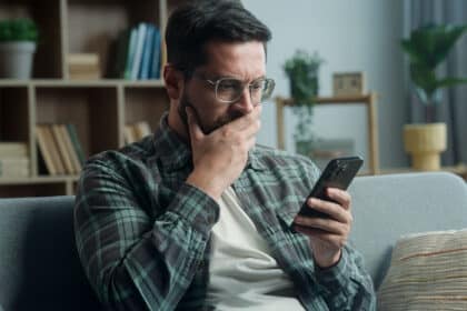 Ein Mann sitzt daheim auf dem Sofa und schaut schockiert auf sein Handy.