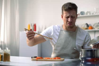 Mann kocht in der Küche und schaut angeekelt, während er sein Essen probiert.