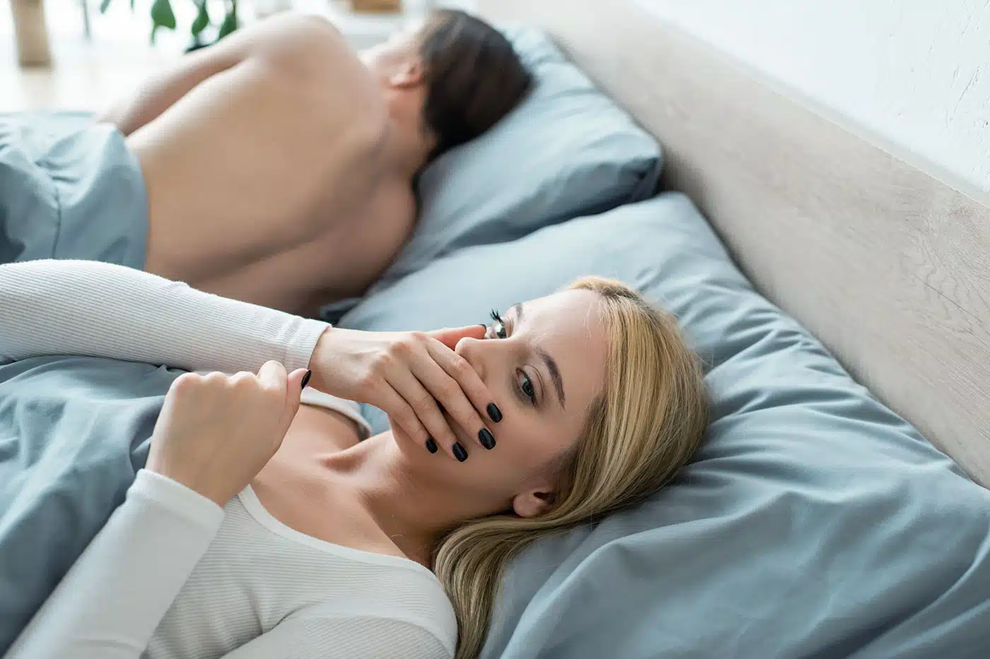 Frau hält sich die Hand vor den Mund, während ein Mann neben ihr im Bett schläft.