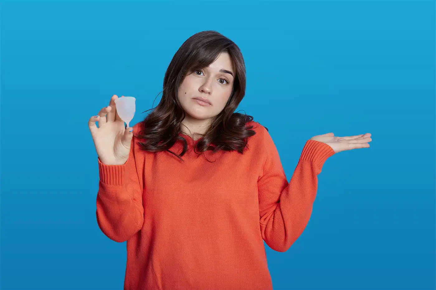 Junge Frau hält eine Menstruationstasse in der Hand und schaut fragend, während sie die andere Hand ratlos hochhält.