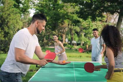 Eine Gruppe von Freunden spielen zusammen Tischtennis im Park und stehen im Kreis um den Tisch herum.