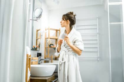 Eine Frau im weißen Bademantel steht in einem hellen Badezimmer und blickt in einen beleuchteten Kosmetikspiegel.