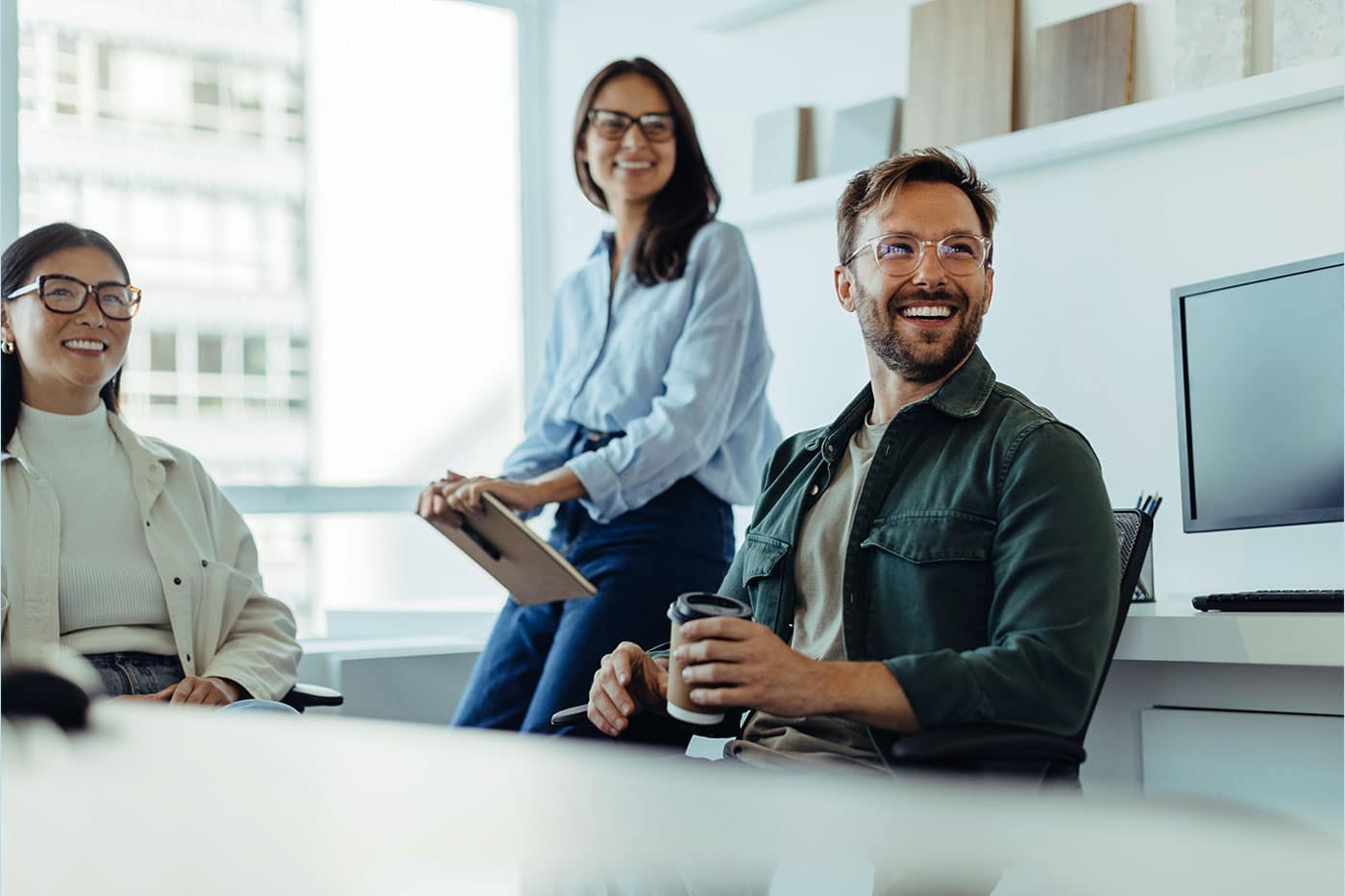 Drei lächelnde Kollegen, zwei Frauen und ein Mann, sitzen und stehen in einem hellen, modernen Büro zusammen.