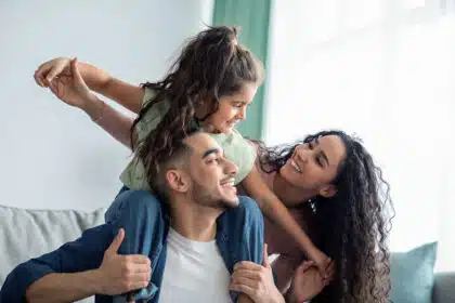 Eine glückliche Familie lacht gemeinsam auf dem Sofa, der Vater trägt seine lachende Tochter auf den Schultern.