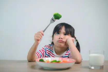 Ein kleines Mädchen hält einen Brokkoli auf der Gabel und schaut ihr essen kritisch an.
